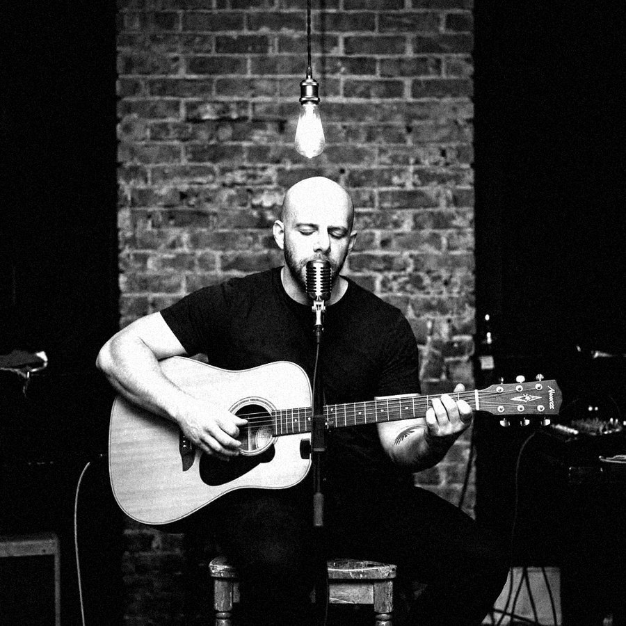 Pete seated with an acoustic guitar under a bare bulb against a brick wall